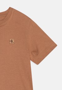 T-shirt marron à manches courtes avec un tissu texturé subtil, un col rond et une étiquette logo carrée en haut à gauche.