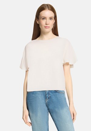 Jeune femme aux cheveux bruns raides portant un haut blanc ample à manches courtes et un jean bleu, sur un fond uni.