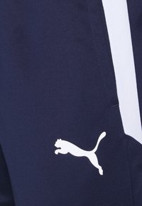 Námořnické sportovní kalhoty s bílými bočními pruhy a velkým bílým logem Puma. Vyrobené z hladké, elastické tkaniny. Mají přiléhavý střih.