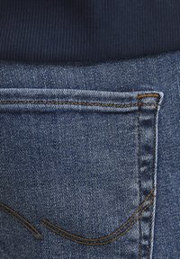 Denimstoff in einem mittleren Blauton mit einer hinteren Tasche, die über eine subtile, geschwungene Sticknaht verfügt, sowie einem sandfarbenen Kontraststich entlang der Nähte.