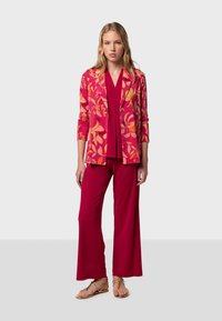 Blazer à motif floral en rose vif et orange, associé à un haut uni rose et un pantalon large. Sandales à fines brides.
