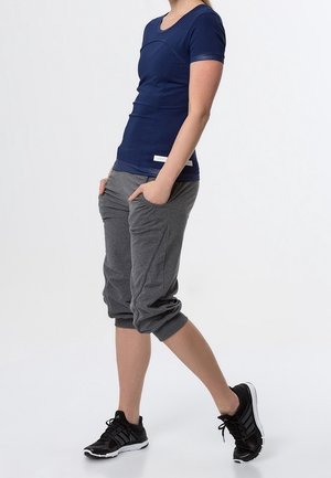 Personne debout les mains dans les poches portant un t-shirt bleu marine, un pantalon gris raccourci et des chaussures de sport noires devant un fond uni.