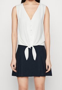 Top bianco senza maniche con scollatura a V profonda, chiusura a bottoni davanti e dettaglio di nodo in vita, abbinato a una gonna plissettata blu navy.