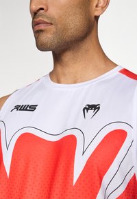 Débardeur sportif blanc avec un design graphique rouge et des accents noirs. Présente un logo sur la poitrine et un motif texturé sur la moitié inférieure.
