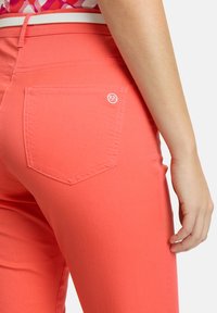 Jean corail avec une poche arrière ornée d'un logo décoratif, texture lisse et design ajusté. Surpiqûres blanches le long des coutures.