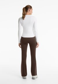 Top alb cu mâneci lungi, ajustat, asortat cu pantaloni maro evazați, textură netedă, design minimalist și decolteu simplu. Încălțăminte sunt adidași de culoare deschisă.