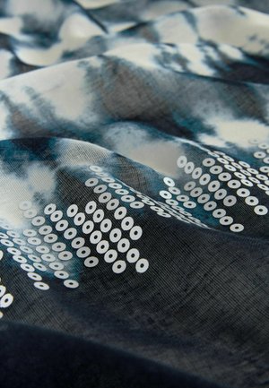 Textília s modro-bielym vzorom batikovania, s bielymi kruhovými flitrami usporiadanými do geometrických tvarov po povrchu látky.