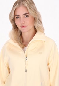 Jauna moteris su banguotais šviesiais plaukais dėvi švelnią geltoną striukę, pozavo prieš šviesų foną, su neutralia veido išraiška.