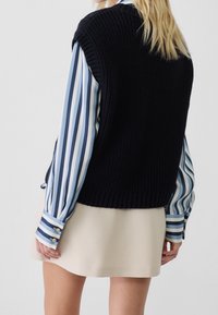 En blond person bär en marinblå stickad ärmlös väst över en blå och vit vertikalt randig långärmad skjorta och en beige kjol, vänd bort.