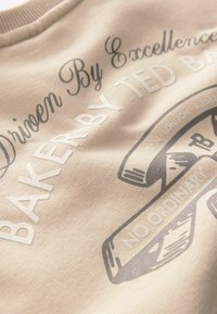 Beige sweatshirt med tryckt text och grafiska designer i mörkare nyanser. Texten inkluderar "Driven By Excellence" och "Baker By Ted Baker."