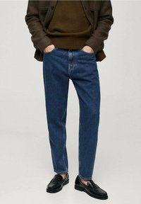 Vaqueros de mezclilla azul oscuro con un corte relajado, que cuentan con cierre de botón y diseño de cinco bolsillos, combinados con mocasines negros y un suéter marrón.