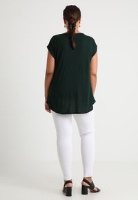 Top verde oscuro de manga corta con dobladillo redondeado, combinado con pantalones blancos. El conjunto tiene una textura suave y un corte relajado. Sandalias de tacón negras visibles.