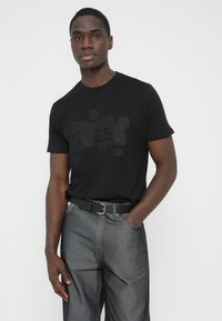 Μαύρο βαμβακερό μπλουζάκι με τη λέξη "GUESS" σε τονικό τύπωμα. Συνδυάζεται με γκρι τζιν παντελόνια και μαύρη ζώνη. Απλός και καθημερινός σχεδιασμός.