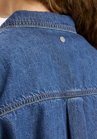 Jeansjacke mit einer hellblauen Waschung, die sichtbare Nähte, einen gefältelten Rücken und einen metallischen Logoschnellverschluss am Kragen aufweist.