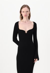 Abito lungo a maniche lunghe nero a coste con scollo a cuore e un accento dorato a forma di cuore al centro. Silhouette aderente, tessuto testurizzato.
