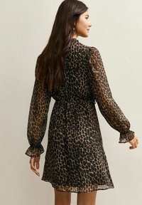 Mujer con cabello largo y oscuro, vestida con un vestido translúcido con estampado de leopardo hasta la rodilla, manga larga y puños con volantes, de perfil frente a una pared lisa.