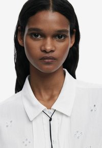 Camicia bianca con tessuto testurizzato, caratterizzata da piccoli disegni floreali ricamati e un accento in corda nera sul collo.