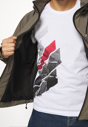 Olive green, zippered jacket worn over a white T-shirt featuring a black and red geometric graphic design. Blue denim jeans visible.