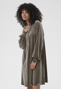 Frau mit lockigem Haar, die ein lockeres, taubengraues Samtkleid mit langen Ärmeln trägt und vor einem schlichten hellgrauen Hintergrund steht.