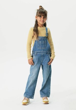 Salopette en denim avec bretelles réglables, dotée d'une poche avant, portée sur une chemise à manches longues à rayures jaunes et blanches. Baskets blanches.