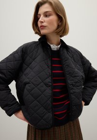 Giovane donna con capelli corti, che indossa un giubbotto imbottito nero, un maglione blu navy con strisce rosse, una camicia bianca colletto e una gonna a fantasia.