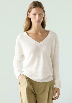 Donna con capelli castano chiaro lunghi, indossa un maglione bianco a V e pantaloni beige, con una mano in tasca, sfondo neutro.