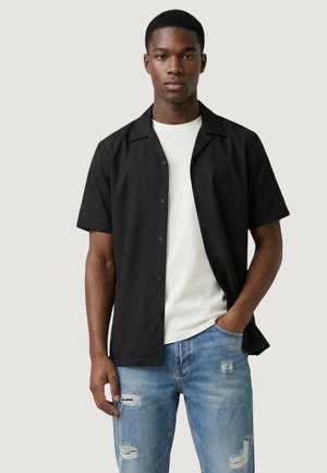Jeune homme portant une chemise noire à manches courtes boutonnée par-dessus un t-shirt blanc, avec un jean bleu clair déchiré, debout devant un fond uni.