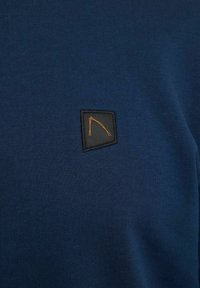 Tmavomodrý pletený sveter so štvorcovou čiernou nášivkou s oranžovým geometrickým dizajnom na ľavej strane. Hladká textúra a priliehavý strih.