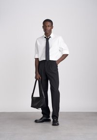 Man standing wearing white shirt with rolled sleeves, black tie, black trousers, and shoes, holding a black handbag against plain background.