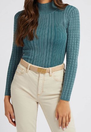 Kvinde iført tealfarvet ribstrikket og kabelstrikket långærmet top med cremefarvede højtaljede jeans og beige bælte med guld "GUESS" spænde.