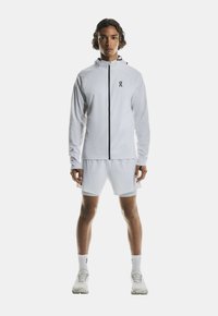 White athletic jacket with black zipper and logo, paired with white shorts. Smooth fabric with a fitted design and hood detail.