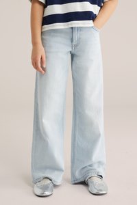 Laps kannab helesiniseid laia säärega teksapükse, hõbedaseid baleriinasussisid valgete sokkidega ning tumesinist ja valget triibulist särki, ühe käega taskus.