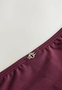 Bordowa tkanina, gładka w fakturze, z okrągłym złotym zapięciem, z marszczonym przeszyciem wzdłuż górnej krawędzi dla dodatkowego detalu.