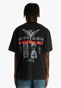 T-shirt nera a maniche corte con una grande stampa grafica sul retro che include un gufo, testo e volti in tonalità di grigio e rosso.