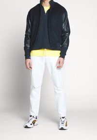 Navy bomberjack met leren mouwen, gelaagd over een donkerblauwe rits, gecombineerd met witte broek en multicolore sneakers.