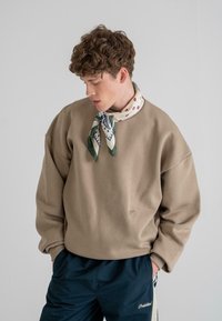 Beige sweatshirt with a relaxed fit, ribbed cuffs, and a small logo detail, paired with a patterned scarf in green and cream tones.