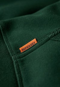 Tessuto verde con una superficie testurizzata, caratterizzato da una piccola etichetta arancione "SUPERDRY" cucita nella cucitura per l'identificazione del marchio.