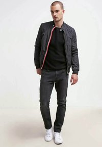 Casaco bomber preto com detalhes em zíper vermelho e branco, usado sobre uma t-shirt preta, combinado com jeans escuros e ténis brancos.
