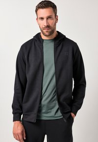 Schwarze Zip-Hoodie mit glatter Textur, ausgestattet mit einer Kapuze und vorderen Taschen, über einem grünen T-Shirt mit dezenten Streifen getragen.