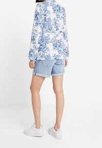 Camicia bianca e blu a fantasia floreale con maniche lunghe, abbinata a short in denim azzurro chiaro e sneakers bianche, con shorts arrotolati.