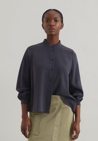 Blusa grigio scuro a maniche lunghe con colletto rialzato e chiusura a bottoni. Presenta una vestibilità ampia e un tessuto testurizzato.