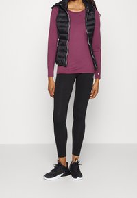 Svart vadderad väst över en långärmad vinröd topp, kombinerad med svarta leggings och svarta träningsskor med vita sulor.