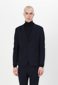 Navy blazer in textured fabric with two front pockets, single button closure, and notch lapels. Worn over a black turtleneck.