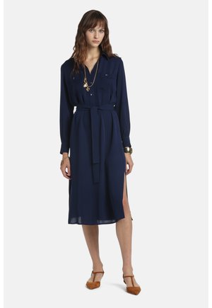 Femme portant une robe midi bleu marine à ceinture, manches longues, poches poitrine, fente latérale, collier doré, bracelets et talons brun à bride en T.
