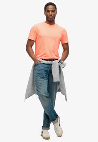 T-shirt arancione neon, jeans di denim blu chiaro, camicia grigia a quadri legata in vita, sneakers bianche con dettagli grigi. Design di un outfit casual.