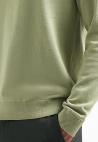 Lichtgroene gebreide trui met een ronde hals en ribgebreide manchetten. Soepele textuur, aansluitend bij de taille; gecombineerd met donkere broeken.