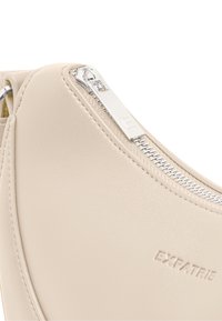 Beigefarbene Ledertasche mit glatter Textur, ausgestattet mit einem silbernen Reißverschluss und dem eingeprägten Markenlogo „EXPATRIE“ an der Seite.