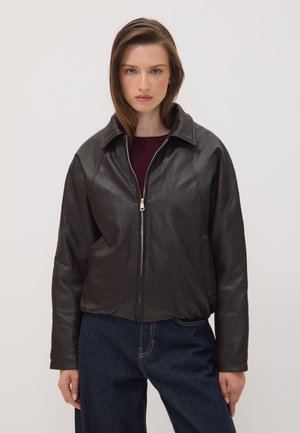 Junge Frau trägt eine schwarze Lederjacke mit Reißverschluss über einem dunkelroten Oberteil und blauen Jeans, steht vor einem einfarbigen hellen Hintergrund.