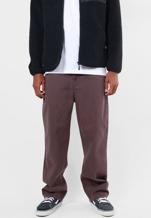 Homme portant un pantalon marron ample, des baskets noires et blanches, une chemise blanche et une veste polaire noire zippée, debout devant un fond blanc.