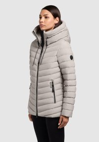 Lys grå pufferjakke med høj krave, vandret quiltet design og to sorte lynlåslommer. Har sorte detaljer og snore.